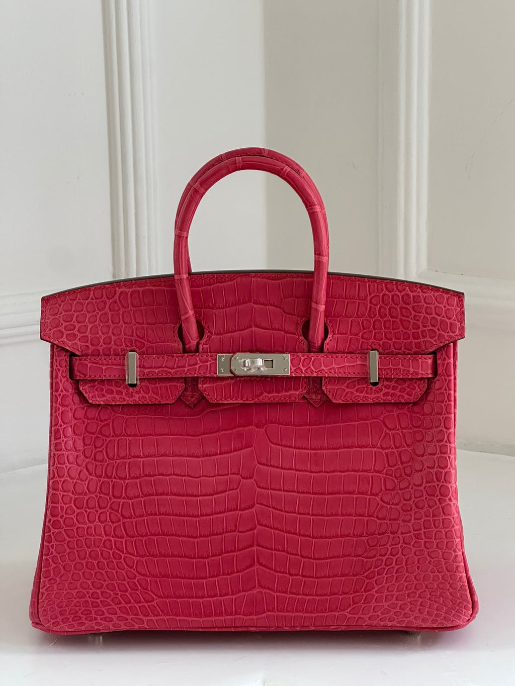 A bright pink Birkin crocodile-embossed leather handbag with silver-tone hardware, displayed against a white paneled background.