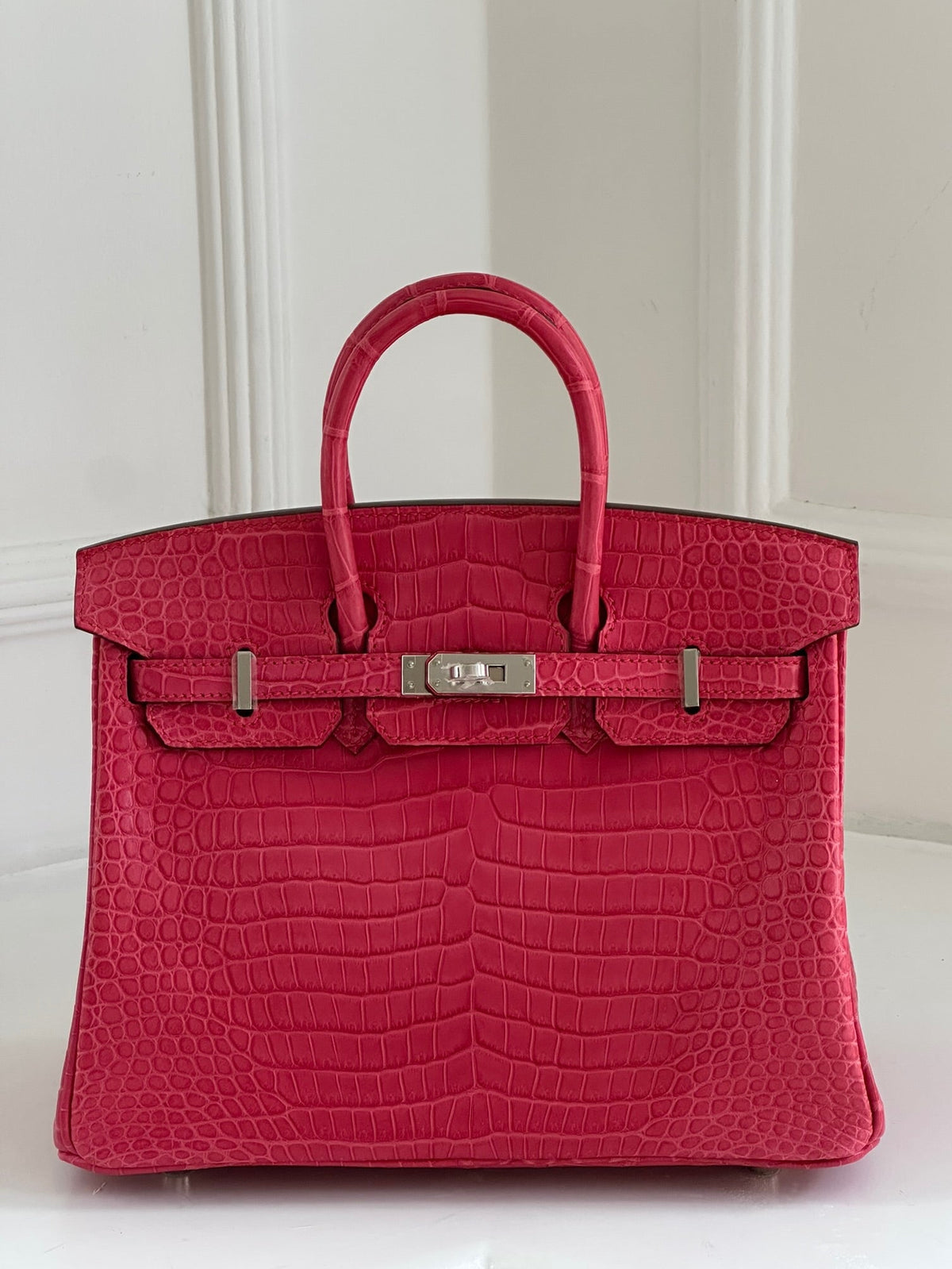 A bright pink Birkin crocodile-embossed leather handbag with silver-tone hardware, displayed against a white paneled background.